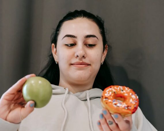 Content young female in hoodie deciding between sweet unhealthy doughnut and green ripe apple in light room